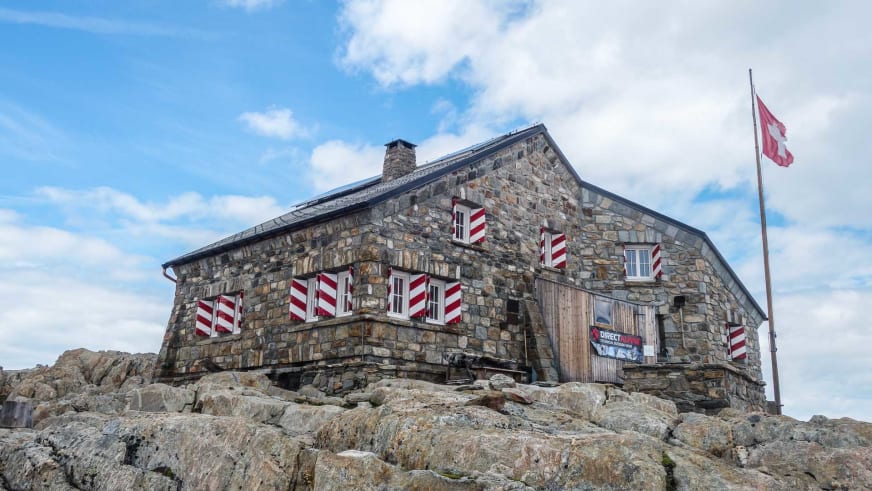 Außenansicht der Tierberglihütte aus graubraunem Naturstein mit rot-weißen Fensterläden auf Felsplateau.