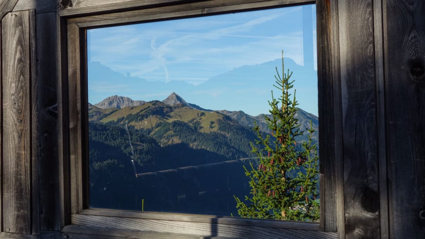 Blick durch das Holzfenster vom Gimpelhaus auf die alpine Berglandschaft mit bewaldeten Hängen und Felsgipfeln.