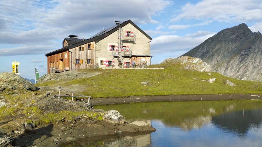 Die Sudetendeutsche Hütte spiegelt sich in der ruhigen Wasseroberfläche eines kleinen Bergsees.