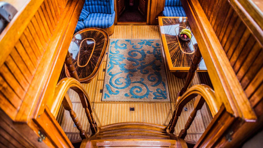 Blick von oben auf die Innentreppe der Ylva mit hellem Holzgeländer, ornamentalem Teppich und blau-weiß gestreiften Sitzkissen.