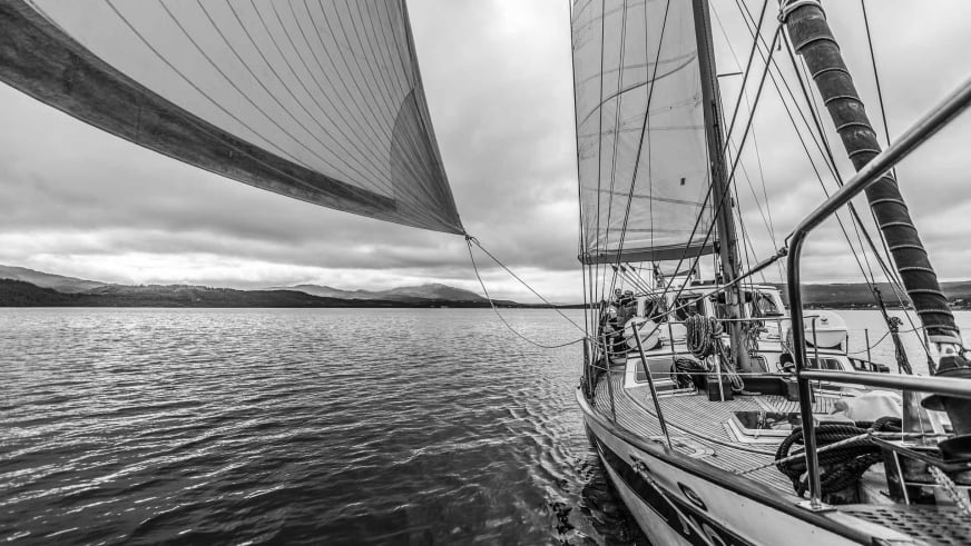Die Ylva segelt mit gesetzten Segeln durch norwegische Gewässer, mehrere Personen auf dem Holzdeck bei bewölktem Himmel.