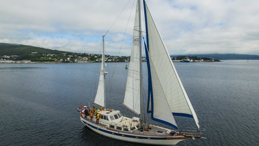 Luftaufnahme der Ylva mit weißem Rumpf und gesetzten Segeln vor bewaldeter norwegischer Küste mit Ortschaften am Ufer.