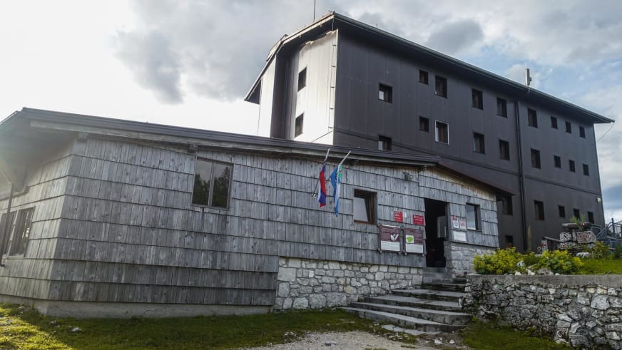 Außenansicht der Berghütte Dom na Komni mit Holzschindelfassade und modernem Betonanbau auf 1520 m in Slowenien.
