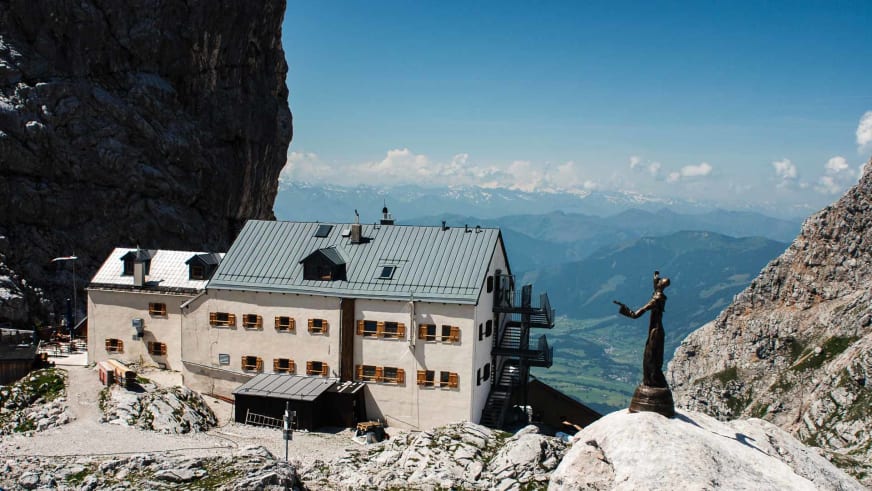 Das Riemannhaus thront auf einem felsigen Plateau in steiler Kalksteinwand mit Wanderer im Vordergrund und weitem Bergpanorama.