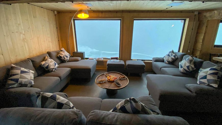 Gemütlicher Aufenthaltsraum der Berghütte Meria Keli mit Sitzgruppe und Bergblick durch große Fenster.