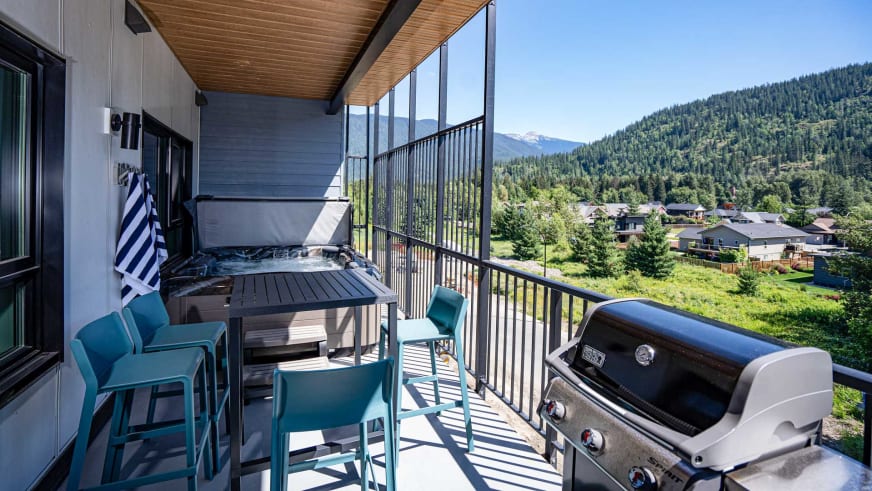 Überdachte Terrasse im Stoked Penthouse Revelstoke mit Holzdeck, Gasgrill und Blick auf bewaldetes Tal.