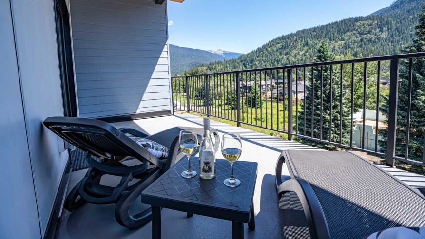 Balkon im Stoked Penthouse Revelstoke mit schwarzen Stühlen, Weingläsern und Blick über bewaldetes Tal.