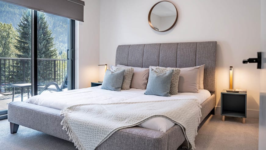 Schlafzimmer im Stoked Penthouse Revelstoke mit Doppelbett und Balkontür mit Blick auf bewaldete Berghänge.