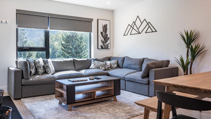 Wohnbereich im Stoked Penthouse Revelstoke mit L-förmiger Eckcouch, Couchtisch und Blick auf Baumkronen.