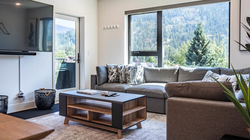 Wohnbereich im Stoked Penthouse Revelstoke mit grauem Ecksofa und Ausblick auf bewaldete Berghänge.