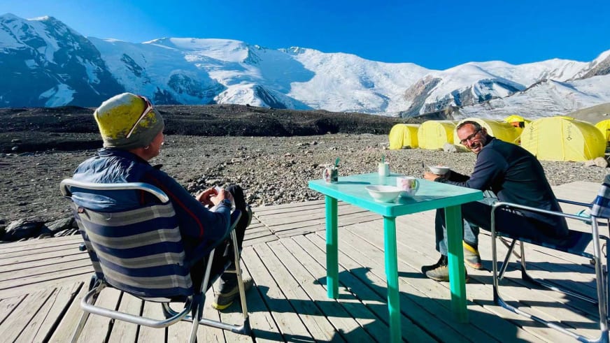 Expeditionsteilnehmer beim Essen am türkisen Tisch im Advanced Base Camp ABC Pik Lenin vor schneebedeckten Gipfeln.