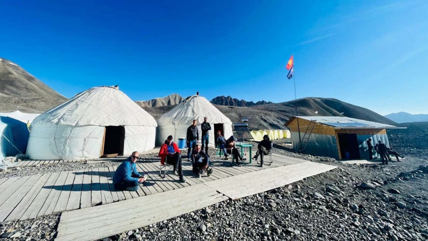 Das Advanced Base Camp ABC Pik Lenin zeigt weiße Jurten auf Holzplattform mit Expeditionsteilnehmern in karger Berglandschaft.