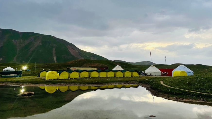 Gelbe Expeditionszelte des Basecamp Pik Lenin spiegeln sich in einem Wassertümpel der kirgisischen Hochebene bei Abendlicht.