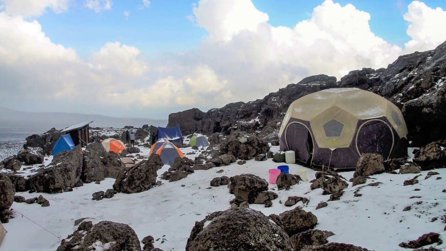 Bunte Expeditionszelte im verschneiten Hochlager des Zeltcamp am Kilimanjaro zwischen vulkanischen Felsblöcken.