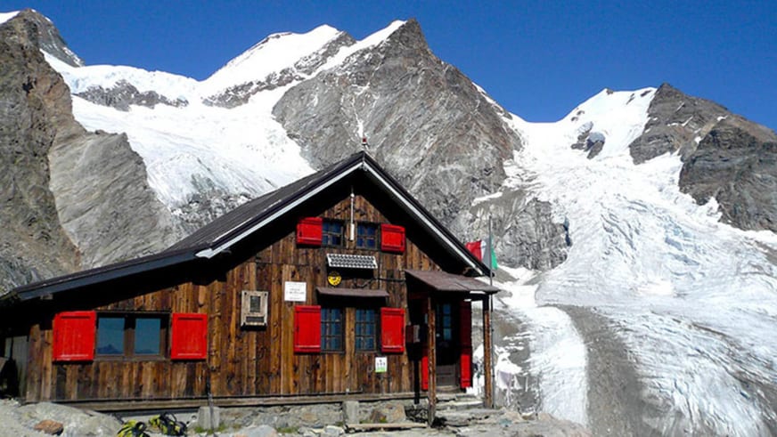 Das Rifugio Ottorino Mezzalama steht als Holzbau mit roten Fensterläden vor einem großen Gletscher in den Walliser Alpen