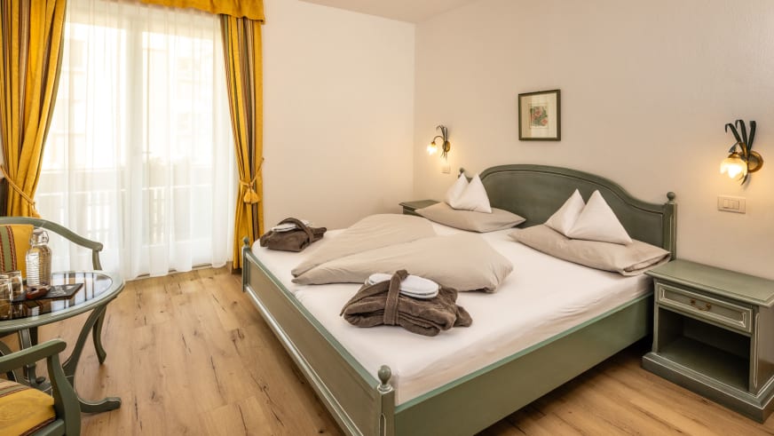 Doppelzimmer im Garni Zimmerhofer Ahrntal mit grünem Holzbett, Glastisch und gelben Vorhängen im traditionellen Alpenstil.