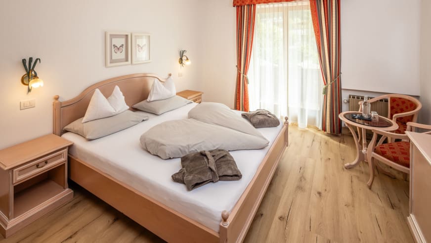 Hotelzimmer im Garni Zimmerhofer Ahrntal mit hellem Holzbett, gestreiften Vorhängen und Hirschgeweih-Wandleuchte im Alpenstil.