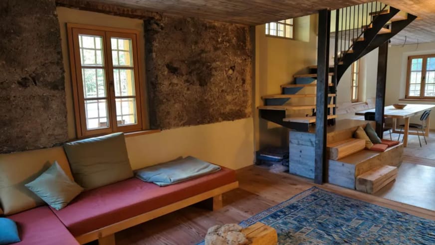 Gemütlicher Wohnbereich der ANAS126 Lodge mit Holz-Wendeltreppe, Natursteinwand und roter Sitzbank mit Kissen.