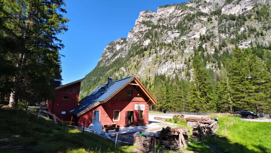 Die rote ANAS126 Lodge mit Satteldach und Solarpaneelen steht vor einer steilen Felswand in den Dolomiten.