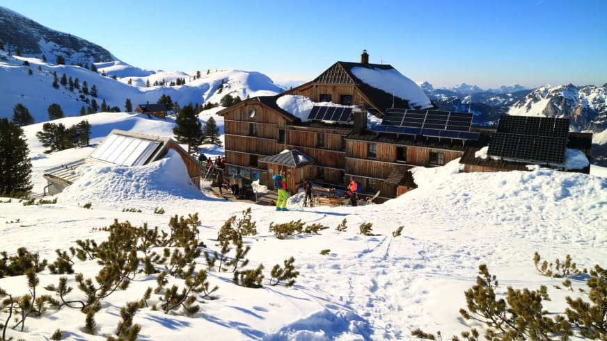 Das verschneite Wiesberghaus auf 1.884 m mit Solarpanelen auf dem Dach und Skifahrern auf der Terrasse im Dachsteinmassiv.