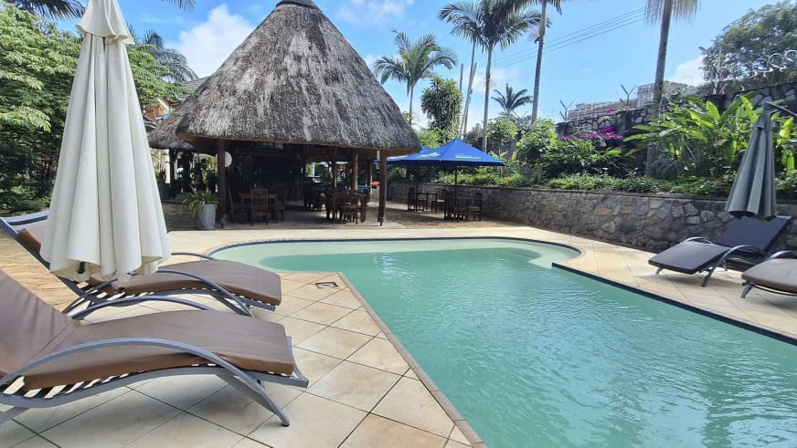 Poolbereich des Entebbe Palm Hotel mit nierenförmigem Schwimmbecken und traditionellem Strohdach-Pavillon inmitten tropischer Vegetation.