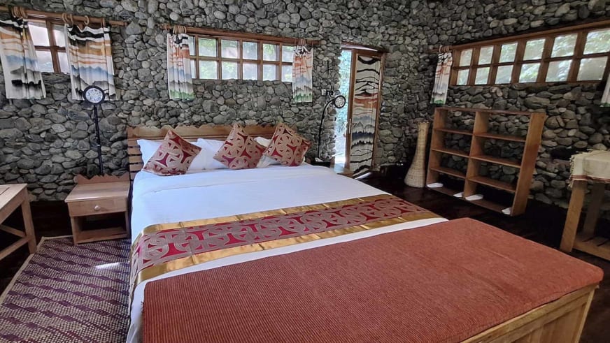 Doppelzimmer der Snow Heights Lodge mit Bruchsteinwänden, traditionellem Bettläufer und Blick durch Sprossenfenster ins Grüne.