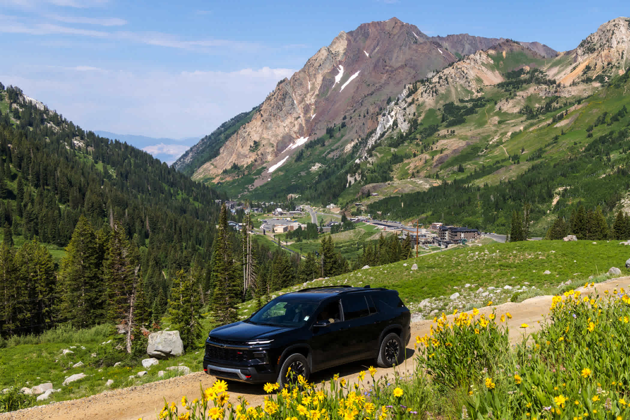 A large, black SUV drives along the unpaved summer road surrounded by wildflowers in the mountains of Alta
