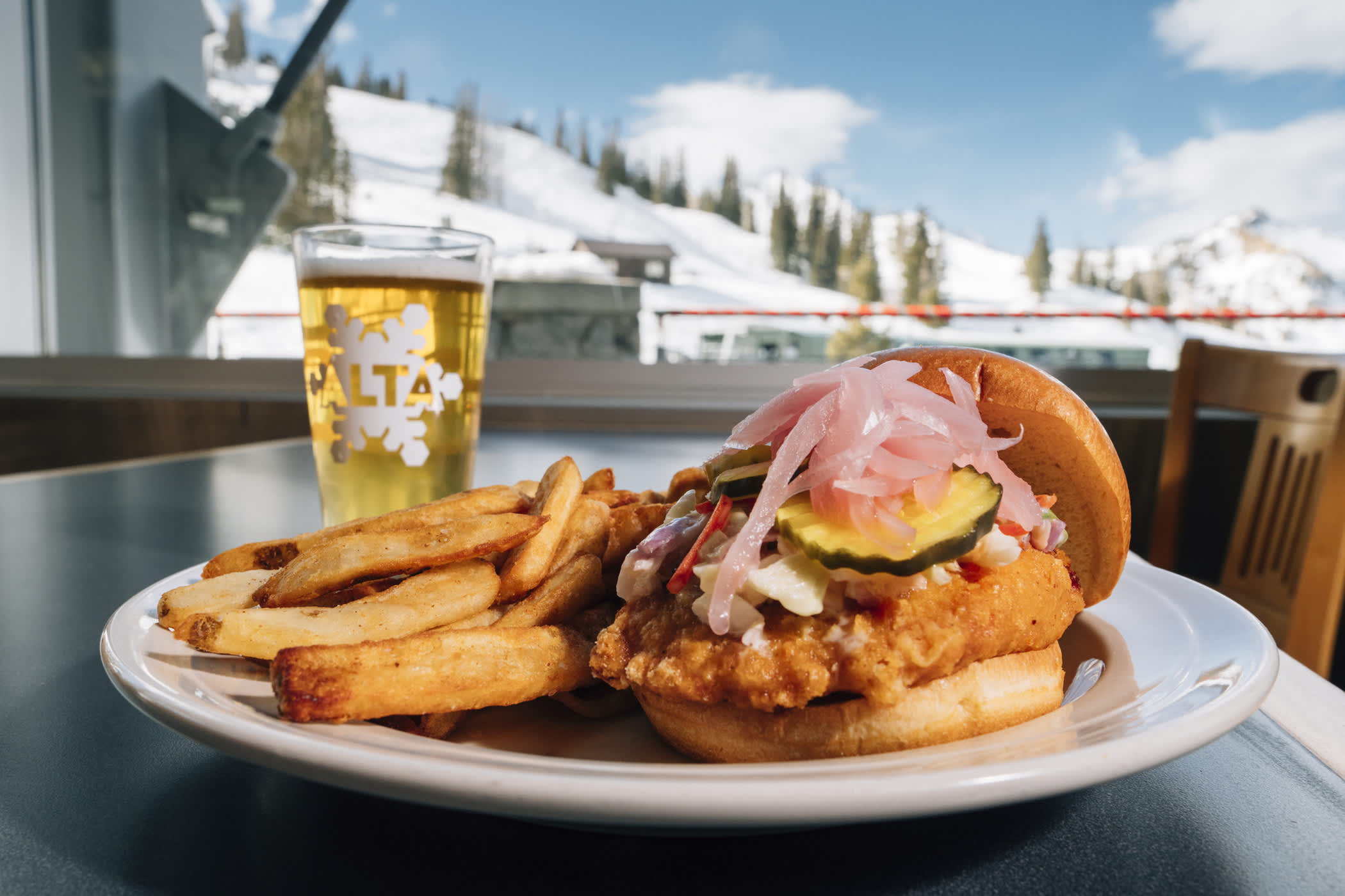 A delicious hamber sits on a table near a pint of beer with a mountain view out the window