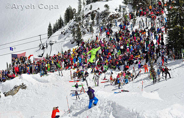 Skier jumping above a crowd of bright colored skiers with alta and American flag in background