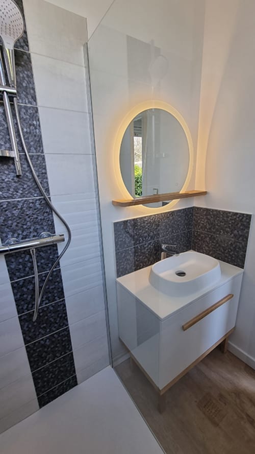 Modern bathroom in Ô LOCH apartment with white vanity, illuminated oval mirror, dark tile backsplash, and walk-in shower at