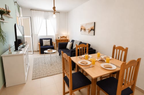 Luminoso comedor con mesa de madera, sillas negras y zona de estar en Apartamentos El Calón Playa
