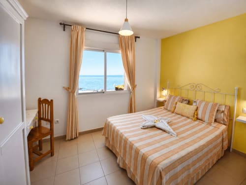 Habitación luminosa frente al mar con pared amarilla, ropa de cama a rayas y vista al océano en Apartamentos El Calón Playa