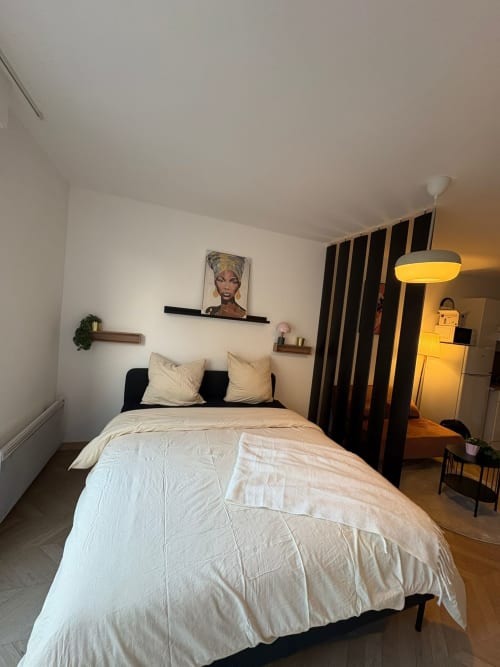 Studio moderne Le Paradis Des Logis : lit avec linge de lit crème, cloison noire, étagères murales et luminaire suspendu