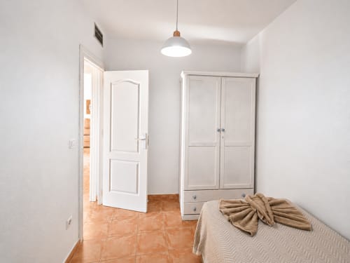 Bedroom with terracotta tile floor, white wardrobe, bed with beige linens, and pendant light at Apartamentos El Calón Playa