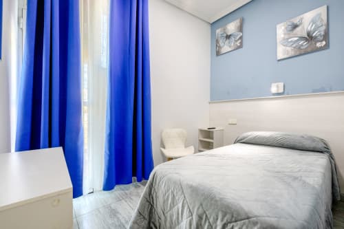 Habitación moderna del Hostal El Molino en Collado Villalba con cortinas azules, ropa de cama gris y vistas a la ciudad