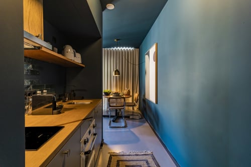 Cozinha compacta moderna em estúdio de hotel no Porto com bancadas em madeira, armários pretos e paredes azuis que se abrem para a área de refeições
