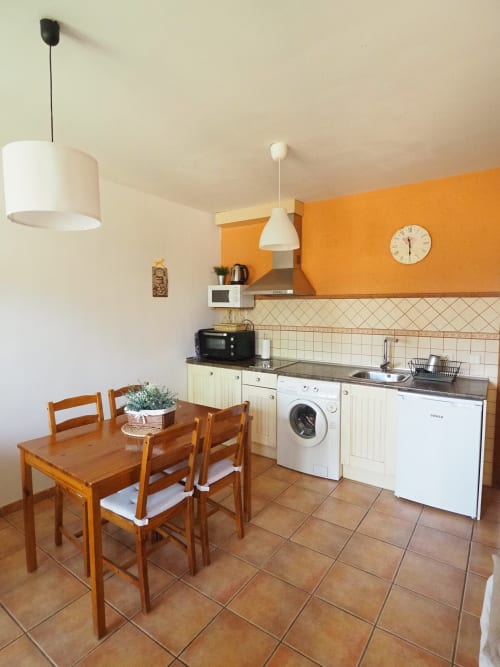 Moderna zona de cocina-comedor con mesa de madera, sillas, electrodomésticos blancos y pared de acento naranja en apartamento rural español