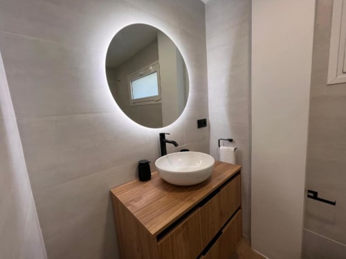 Baño moderno con espejo iluminado redondo, lavabo blanco de cerámica sobre tocador de madera en El HierroStay Apartments La Chalana