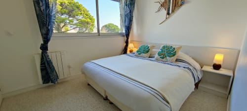 Chambre lumineuse avec vue mer à Mer et Maisons, lit blanc, coussins turquoise, grande fenêtre sur les arbres
