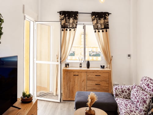 Luminoso salón con tocador de madera, cortinas estampadas, sillón floral y acceso a balcón en Apartamentos El