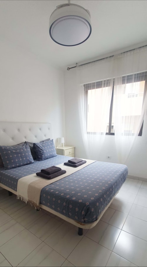 Moderna camera da letto nell'appartamento El Médano con biancheria blu decorata, testiera bianca e luce naturale dalle finestre