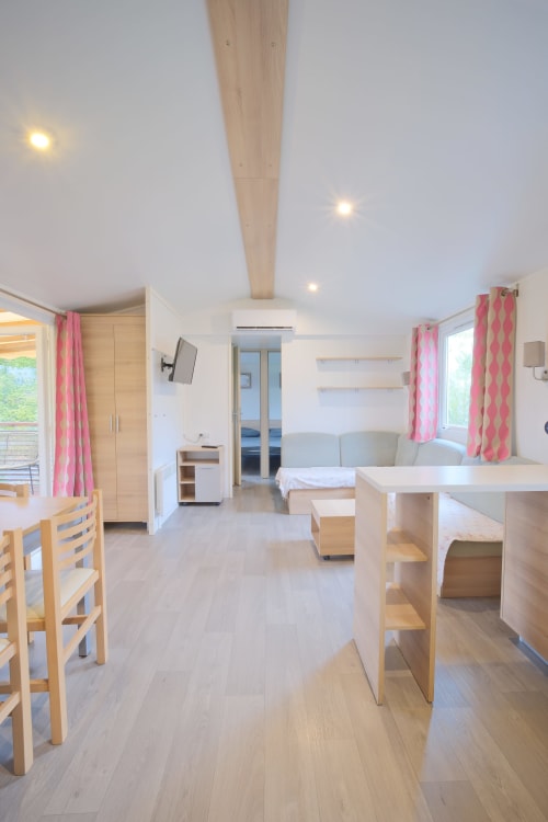 Intérieur lumineux et moderne du mobilhome à Vias Plage avec mobilier en bois, rideaux roses, séjour ouvert et vue sur mer