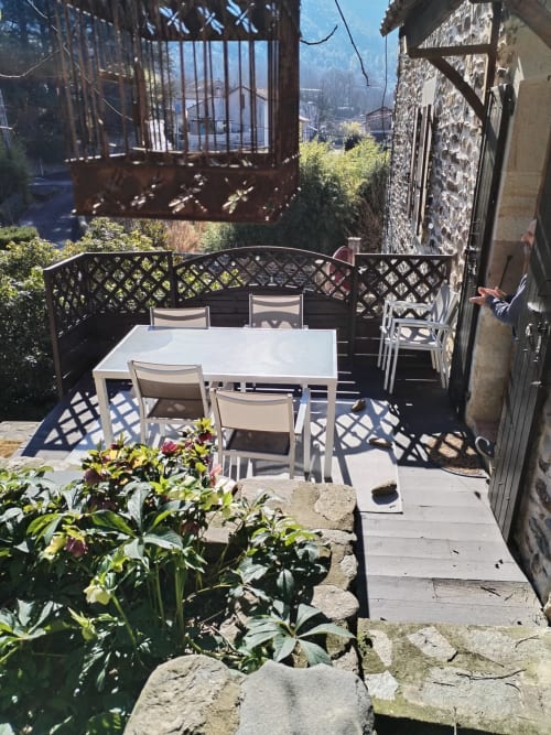 Sunny terrace with white dining table and chairs, mountain views, flowering plants, and rustic stone walls at French gîte
