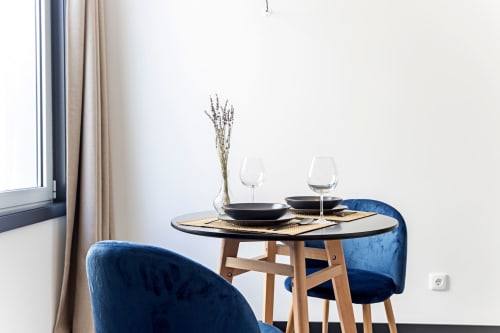 Moderno espaço de refeições em estúdio de hotel no Porto com cadeiras de veludo azul, mesa redonda, talheres e flores secas à janela