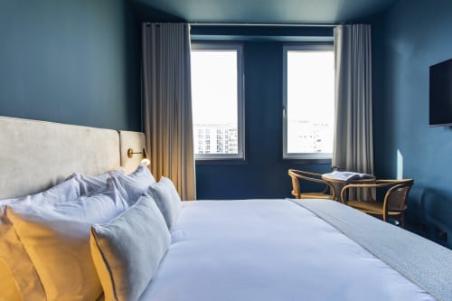 Quarto de hotel moderno no Porto com paredes azuis, roupa de cama branca, grandes janelas com vista para a cidade, secretária e cadeiras de madeira