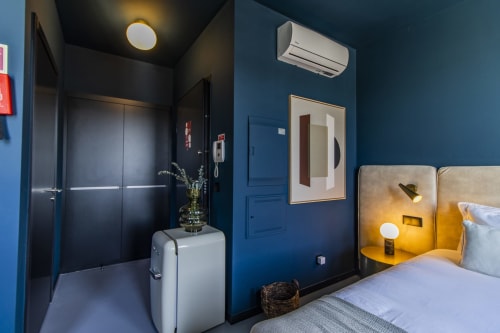 Quarto estúdio compacto em hotel no Porto com paredes azuis, cama com cabeceira dourada, ar condicionado e banheiro moderno