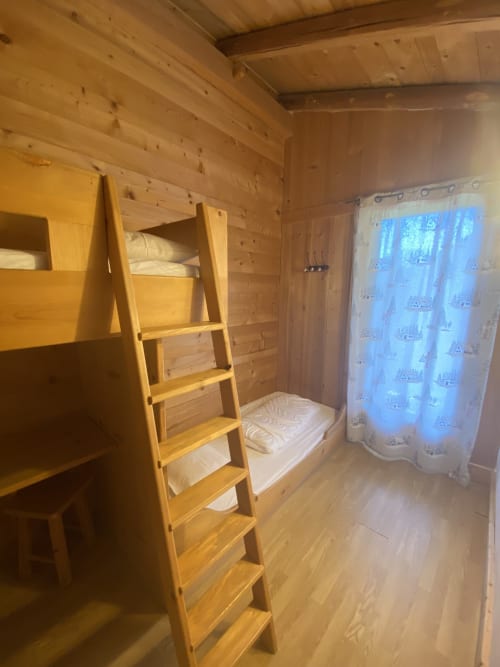 Chambre de montagne avec lit superposé, échelle en bois, literie blanche, fenêtre et rideaux, poutres apparentes