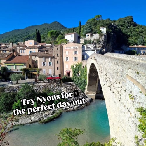 Picturesque stone bridge over turquoise river in Nyons, France, with charming village buildings and mountains.