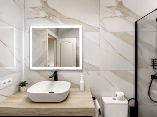 Baño moderno con paredes de mármol, espejo iluminado LED, lavamanos blanco y accesorios negros en Apartamentos El Calón Playa