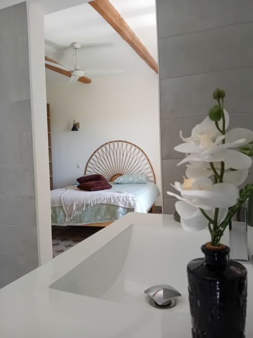 Chambre moderne dans le gîte Chez Laure & Marc avec tête de lit en rotin, poutres apparentes, ventilateur et fleurs blanches sur le bureau